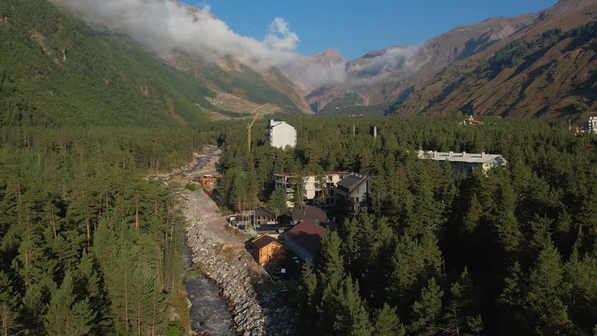 A mountain town nestled in a lush valley with a river running through. Buildings are scattered among the trees, blending with the landscape. Mountains rise in the background.