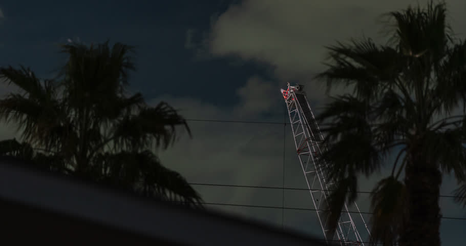 Fever-dream scene with a construction crane and chaotic wind-blown palm trees that tower over a roofing gable at night.