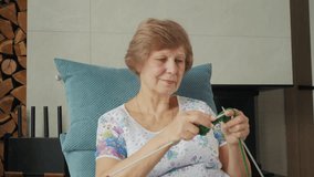 Elderly woman knitting with green yarn in a cozy armchair near a fireplace, focused on her work with a concentrated expression. High quality 4k footage - Powered by Shutterstock - Get 15% off with code: PIKWIZARD15