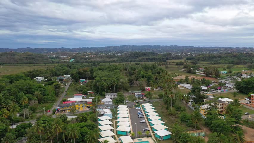 A drone view of a rural coastal landscape featuring gentle waves, green spaces and scattered houses and cabins under cloudy sky in Puerto Rico
