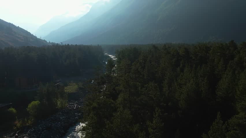 A rushing river meanders through a lush evergreen forest. The towering mountain slopes in the background create a dramatic contrast to the peaceful valley below.