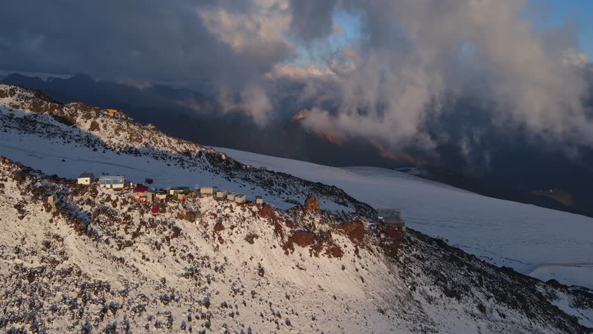 An aerial view of the glacial landscape of Mount Elbrus, featuring deep crevasses and rugged snow-covered slopes. The vast ice fields stretch into the distance under a dramatic sky.