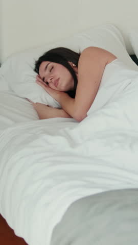 Vertical video, A young woman sleeps soundly in her comfortable bed, enjoying a peaceful rest. The bedroom is simple and cozy, offering a sense of tranquility and relaxation.