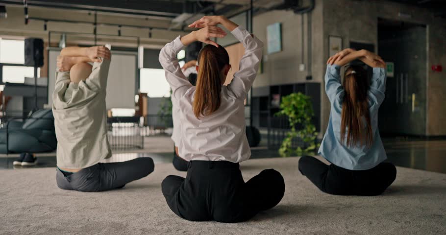 A rear view of office workers meditating and practicing yoga on a carpet focusing on their well-being during a work break in a modern office
