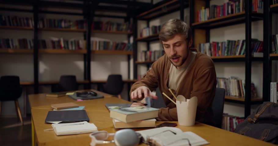 A guy with a mustache and beard sits at a table opens books and takes notes while studying in the library