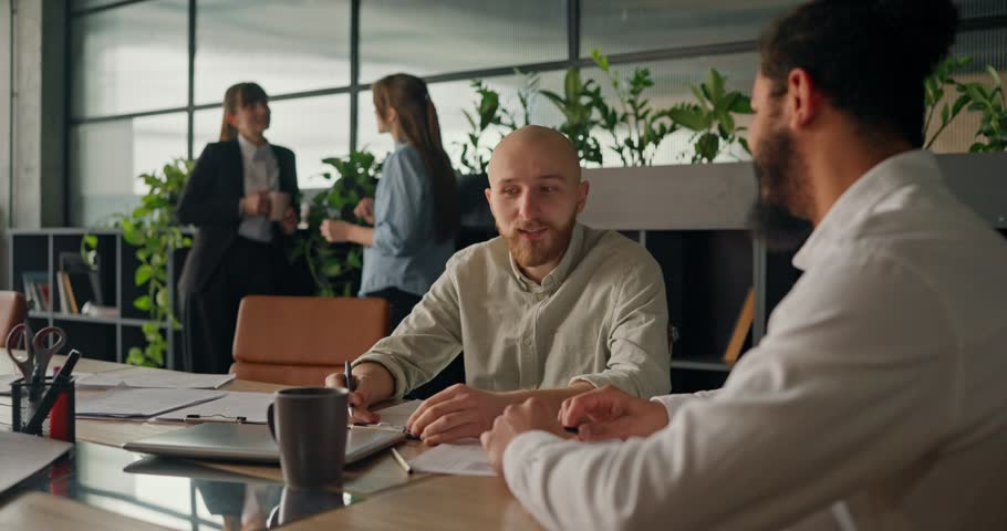 Two office workers sit at the table surrounded by papers and laptops chatting casually and taking a break from work in a modern office