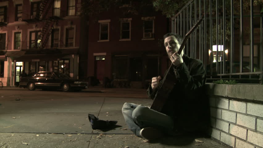 Young man playing a worn-out guitar on the night time streets of Manhattan, NY.