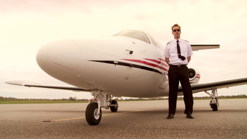 Pilot in uniform and young woman in a black dress pose next to an executive jet