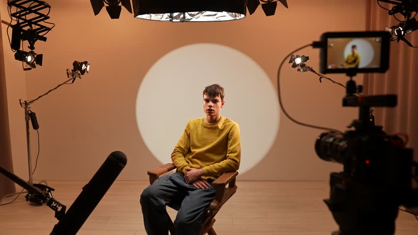 Studio for photo video production filming with lighting equipment and cyclorama, model actor sits in front of camera on chair talking, stylist comes to fix makeup.