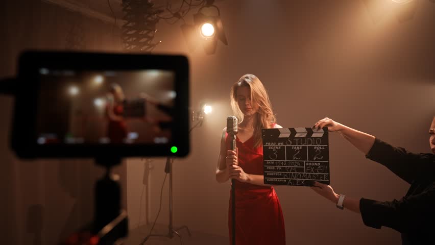 Studio for photo video production filming with lighting equipment, focus on camera display, woman singer in red dress and assistant with clapperboard.