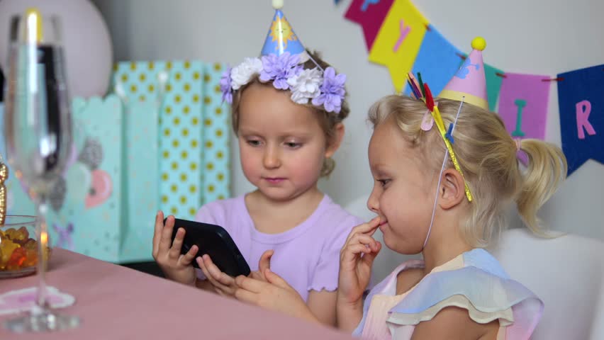 Two little girls wearing party hats are sitting at a table watching something on a smartphone during a birthday party, with gifts and decorations in the background