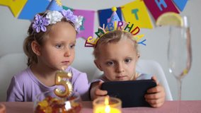 Twin sisters wearing birthday hats are sitting at a table with a lit candle, a glass of water, a bowl of candy and a happy birthday banner in the background, watching something on a smartphone - Powered by Shutterstock - Get 15% off with code: PIKWIZARD15