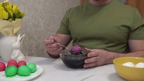 4k. A man shows the process of coloring chicken eggs purple, sitting at a table with a bouquet of yellow tulips and Easter bunny figurines. - Powered by Shutterstock - Get 15% off with code: PIKWIZARD15