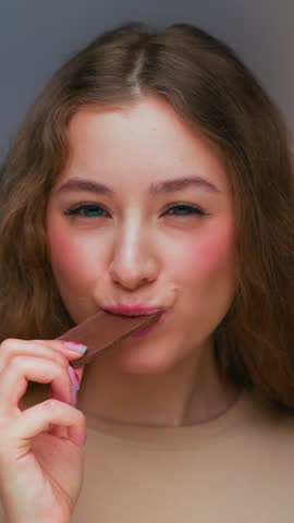 Portrait of Young Woman Eating Sweet Food as Looking at Camera. Real Person on Diet Consumes Chocolate Candy Bar as Lunch Snack. Playful Female Human Inside Cozy Living Room Alone with Sugary Fastfood