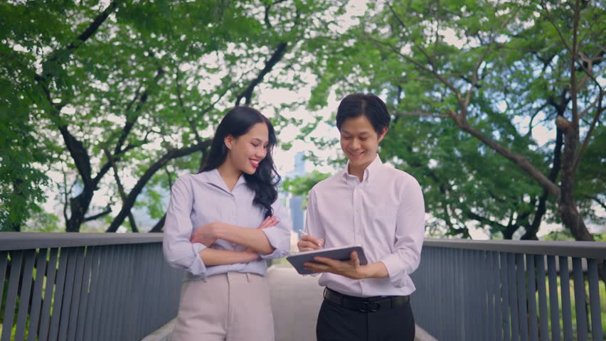 Asian young businessman and woman partner working outdoors in the garden. Attractive colleagues employee feel happy and relax while spend leisure time walking outside office company with confidence.