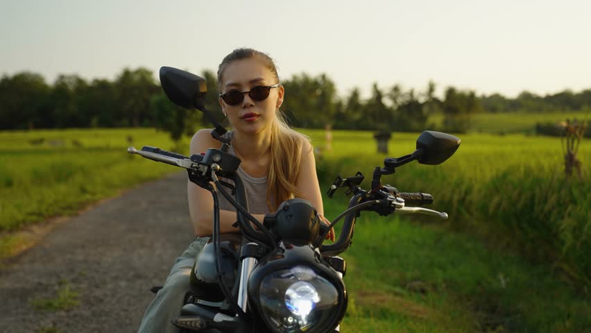 Confident young woman wearing sunglasses leans on a motorcycle, moto driver in a green rural landscape at sunset. Bike driving and moto trip concept