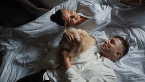 A couple in sleepwear lies on a bed, smiling and petting a fluffy dog, enjoying a cozy and intimate moment together. Loving family at home - Powered by Shutterstock - Get 15% off with code: PIKWIZARD15
