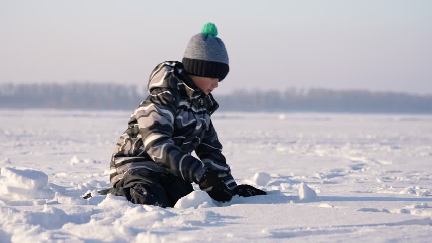 A child is kneeling on a snow-covered frozen lake, engaging in playful activities. The cold winter air is clear and crisp, creating a serene atmosphere around him.