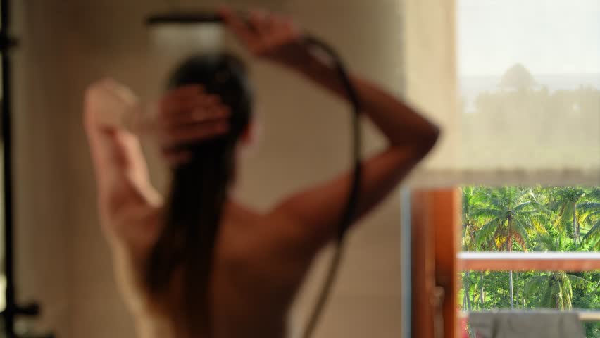 A woman takes a shower, rinsing her hair while facing a window overlooking lush tropical palms. The camera focuses on the serene jungle view. Soft, relaxing atmosphere.