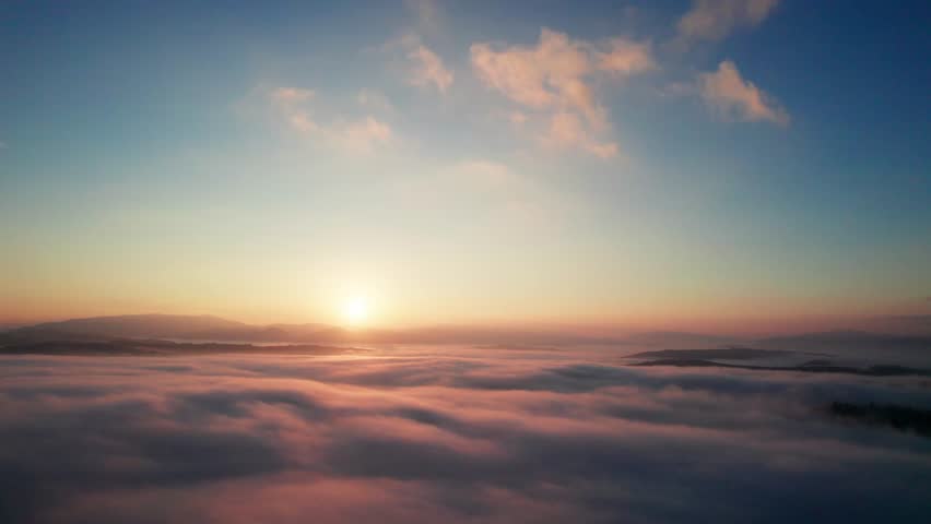 Aerial view of sun rises above sea of clouds, painting sky with soft hues of orange and blue. Light, wispy clouds scatter across horizon, capturing serene and beauty of dawn breaking over landscape.