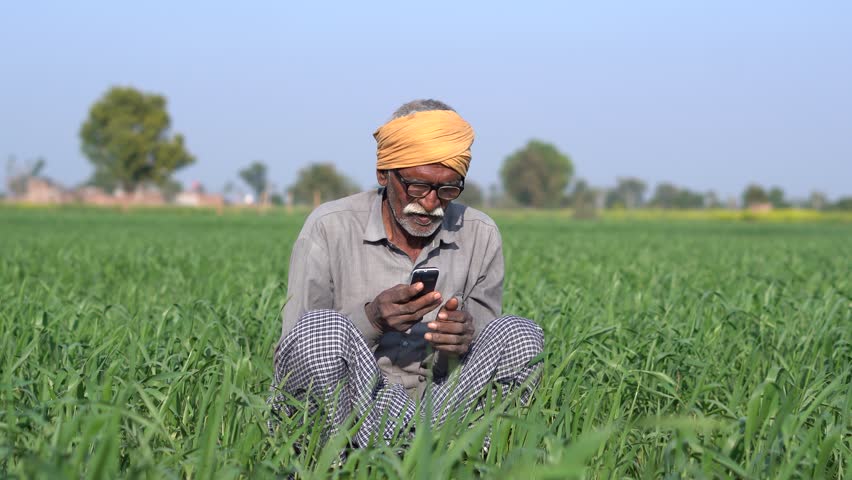 Senior rural indian farmer sitting outdoor at agriculture filed talking on mobile phone. Elderly man using cell phone, Wireless technology. People on india.