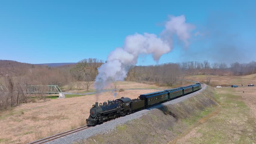 A steam locomotive chugs along winding tracks, releasing puffs of smoke as it passes through open fields and distant hills under a bright blue sky.