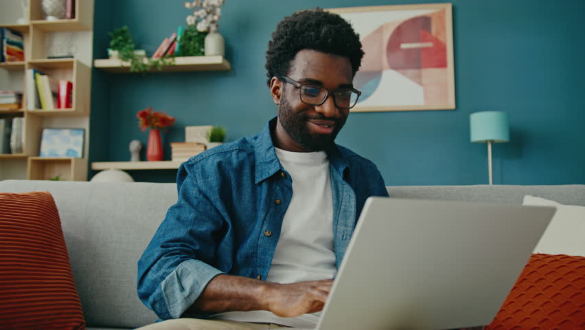 Excited African American man wearing glasses sitting on stylish sofa using laptop. Smiling while waving during video call with relative. Enjoying friendly conversation in cozy modern home.
