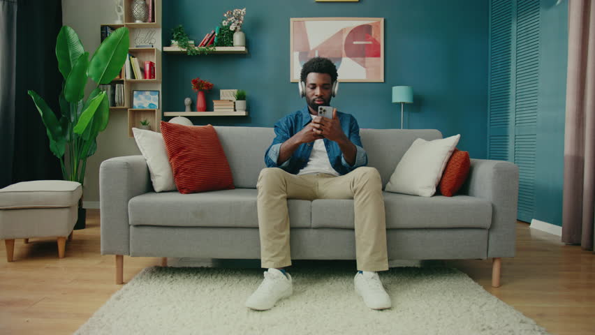 Handsome African American man sitting on comfy sofa wearing headphones and using smartphone. Browsing messages or listening to music while resting. Enjoying peaceful moment in cozy modern home. - Powered by Shutterstock - Get 15% off with code: PIKWIZARD15