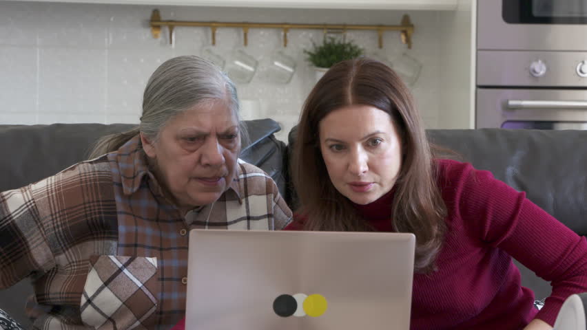 A senior woman and a younger woman are intently focused on a laptop, appearing to struggle with the technology, while sitting on a couch in a home setting.
