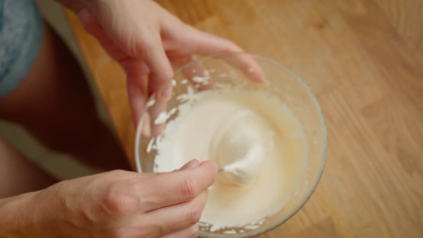 Woman carefully mixing whipped cream in glass bowl using spoon for dessert preparation. Female gently folding creamy mixture to achieve smooth texture during cooking. Lady stirring whipped mixture