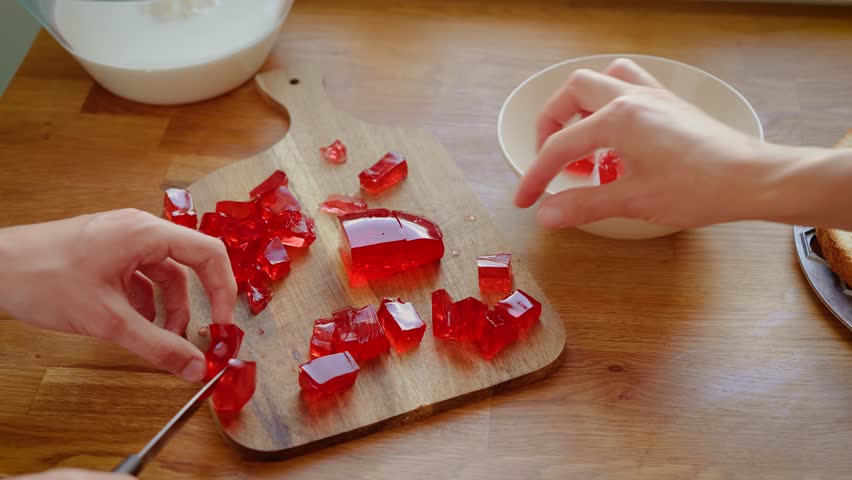 Mother and daughter slicing red jelly into cubes on wooden board. Female adult and girl preparing gelatin pieces for dessert. Lady and child cutting colorful jelly for cake decoration