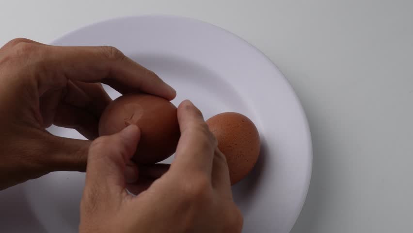 Someone cracks the shell of a boiled egg, peels it, and places it on a white plate.