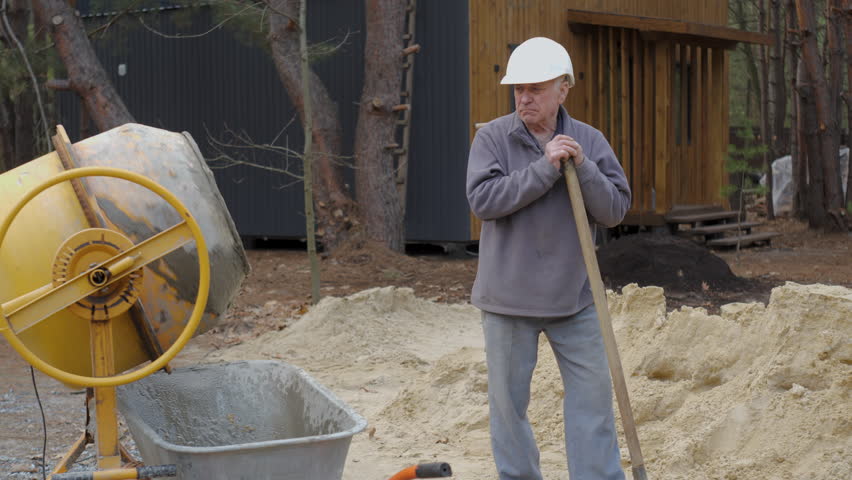 An elderly construction worker wearing a helmet holds a shovel and rests at a construction site near a concrete mixer