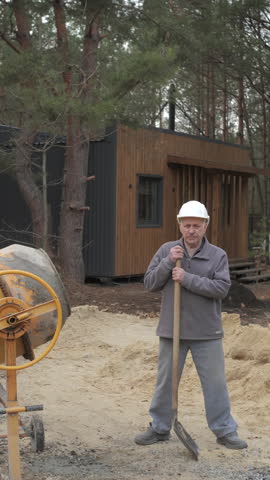 An elderly construction worker wearing a helmet holds a shovel and rests at a construction site near a concrete mixer. Vertical footage