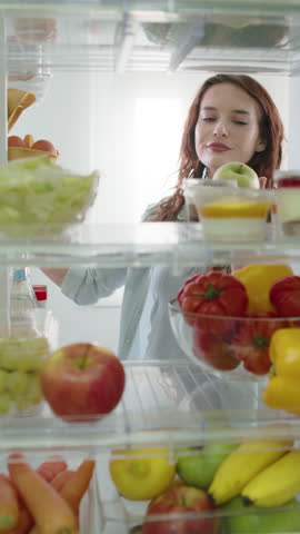 Woman stands in front of open fridge, reluctantly deciding between a dessert and a fresh apple. She chooses the healthy option, making a healthy diet choice. POV shot from inside kitchen refrigerator