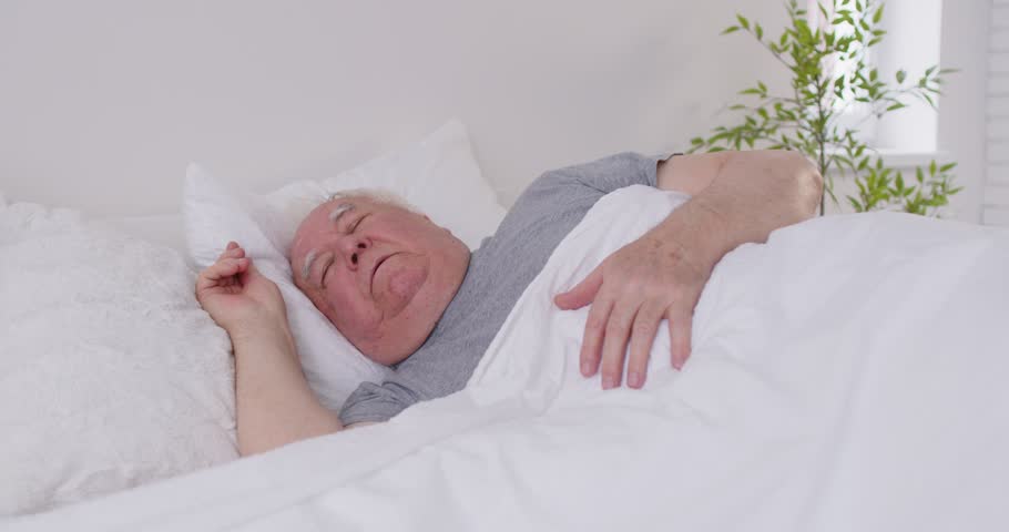 Senior man resting in bed at home, sleeping peacefully in the early morning. The calm environment of bedroom allows him to slumber deeply and dream as part of everyday morning routine.