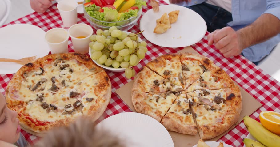 Family eating pizza. Group unrecognizable people having dinner enjoying two takeout or homemade pizzas. Cropped shot hands taking slices from board on red white gingham vichy check pattern table cloth