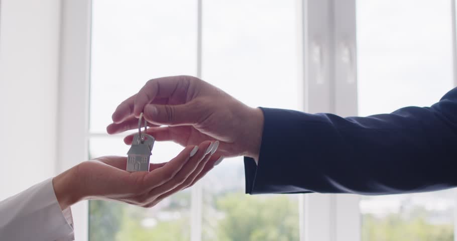 Professional real estate agent gives key to woman who is buying new house or apartment. Close up hand of male realtor giving key to female client, light white window in background, copy space for text