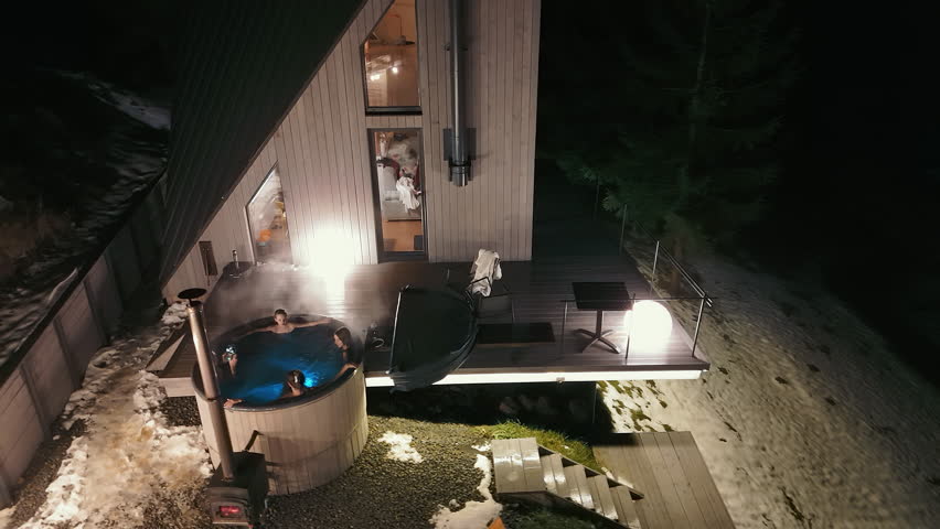 Family enjoying outdoor hot tub at night, beside modern cabin with illuminated deck and snowy surroundings.