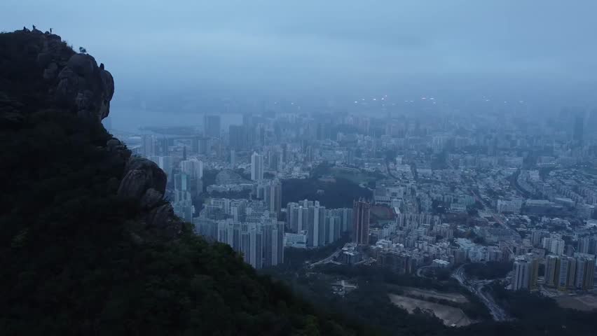 Aerial drone footage flying over and besides a steep rock cliff with plants and reveals the city of Hong Kong in the background that is covered in mist, fog or smog during a cloudy dusk or dawn day.