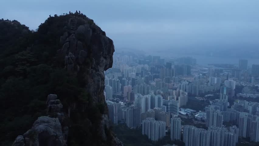 Gorgeous aerial drone footage of the city Hong Kong in China during a misty foggy or smog covered dusk or dawn day with a large mountain ricky cliff in the foreground. Tall building visible in city.