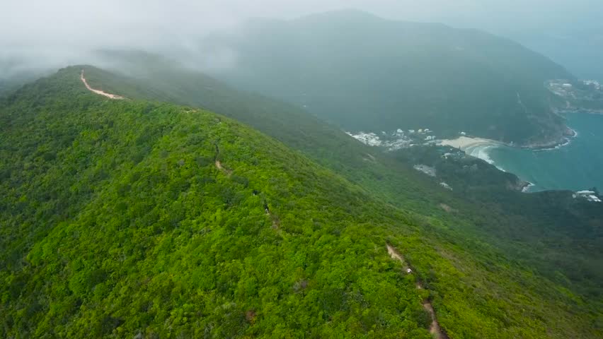 Aerial drone footage flying over Chinas green nature mountains during a sunny day while mountains have white clouds, mist and fog surrounding the peaks. Small sandy beach with ocean visible down below