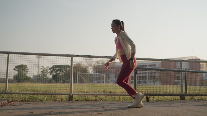 A woman is running on a sidewalk in front of a fence. She is wearing a pink shirt and red pants