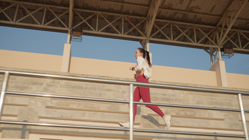 A woman is running on a metal railing. She is wearing red pants and a white shirt. The railing is located on a stadium bleacher