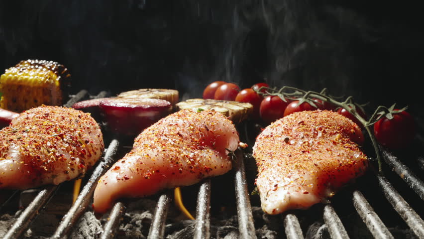 Smooth movement. Chef's hand gently turns juicy chicken steaks sprinkled with seasoning. In background there are delicious ruddy vegetables. Low heat under grill grill perfects this delicious dish.