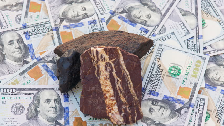 Three gemstones, Tiger Iron, Hematite, and Obsidian, rotate on a turntable atop genuine US 100-dollar bills. Dark stones with metallic and earthy tones contrast with the green currency background.