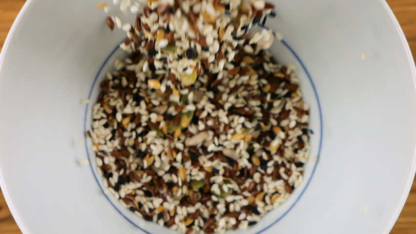 Salad seed mix is ​​poured into a white ceramic bowl. Sesame, flax, caraway, sunflower and pumpkin seeds. Top view. Slow motion.