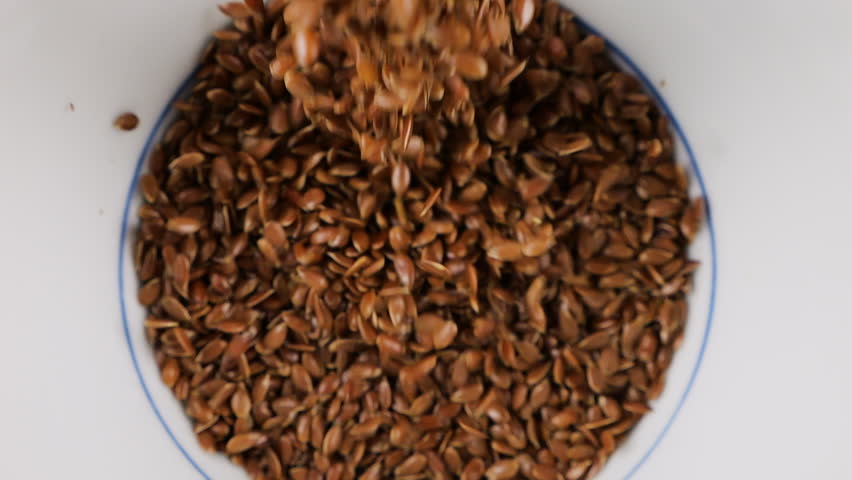 Flax seeds are poured into a white ceramic bowl. Top view. Slow motion.