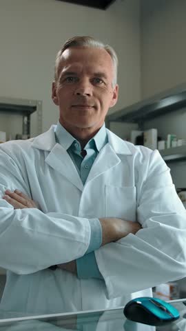 Mature male pharmacist in the interior of a pharmacy. Portrait of a confident man with crossed arms in white robe and shirt looking at the camera with medicines on the shelves in the background