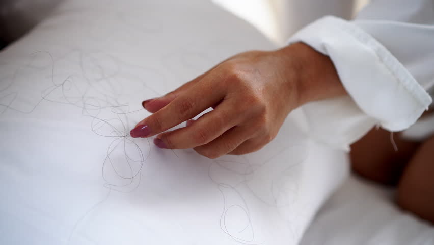 closeup of a woman’s hand cleaning fallen hair from the bed and pillow caused by stress, hormonal changes, or health problems, dealing with hair loss at home